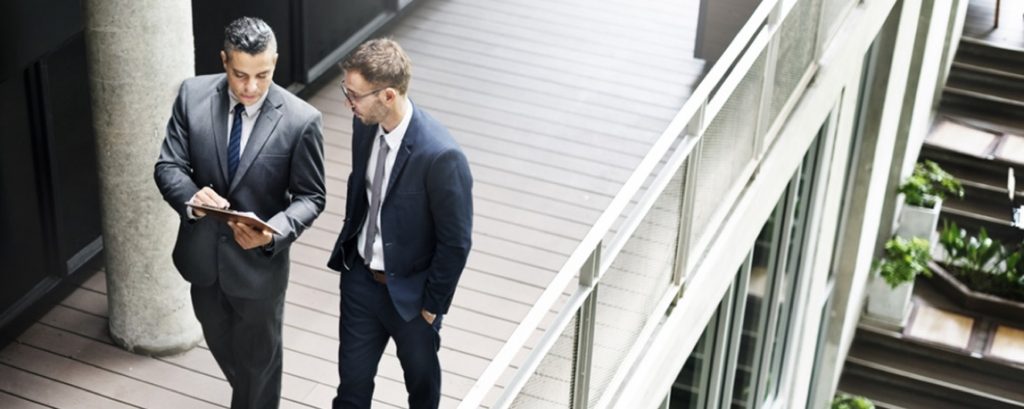 Two businessmen looking at a chart while walking down an exterior walkway of an office building